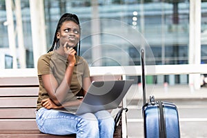 Young african woman with hand luggage in international airport terminal, working on her laptop while waiting for flight