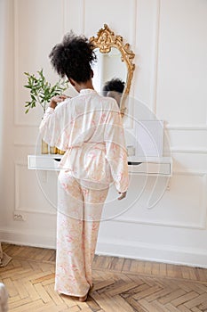 Young african woman in pajamas standing against mirror and looking at her reflection in it