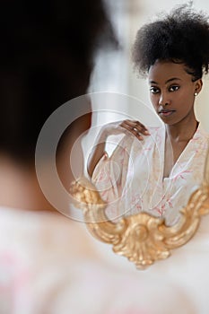 Young african woman in pajamas standing against mirror and looking at her reflection in it