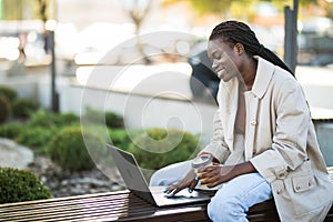 Young african woman in park. Beautiful woman sitting on bench drinking coffee and using laptop via airpods