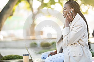 Young african woman in park. Beautiful woman sitting on bench drinking coffee and using laptop via airpods