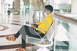 A young African woman in a protective mask sits in a chair in the waiting room, listening to music with headphones and