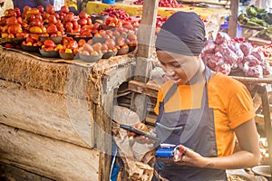 Young african woman selling tomatoes in a local african market using her mobile phone and holding a mobile pos device.