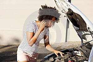 Young african woman standing by broken down car parked on the road and calling for assistance