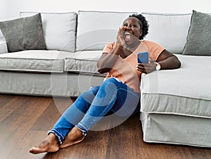 Young african woman using smartphone sitting on the floor at home shouting and screaming loud to side with hand on mouth