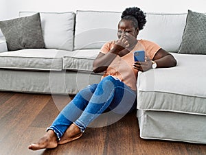 Young african woman using smartphone sitting on the floor at home smelling something stinky and disgusting, intolerable smell,