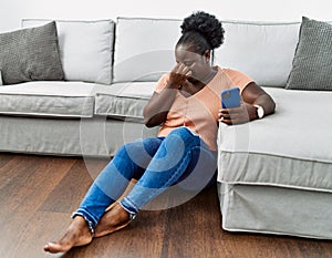 Young african woman using smartphone sitting on the floor at home tired rubbing nose and eyes feeling fatigue and headache