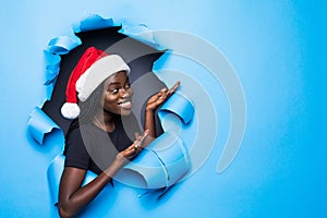 Young african woman wear in santa hat preseting new product on cope space standing in blue torn paper hole background