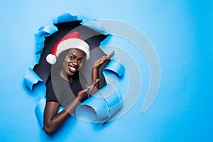 Young african woman wear in santa hat preseting new product on cope space standing in blue torn paper hole background