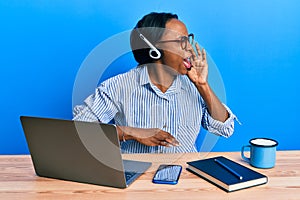 Young african woman wearing call center agent headset shouting and screaming loud to side with hand on mouth