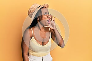 Young african woman wearing summer hat over yellow background shouting and screaming loud to side with hand on mouth