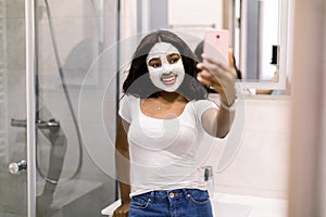 Young African woman with white clay mud facial mask using cellphone in the bathroom, making selfie photo, posing in the