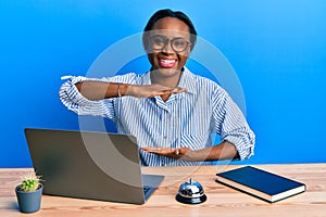 Young african woman working at hotel reception using laptop gesturing with hands showing big and large size sign, measure symbol