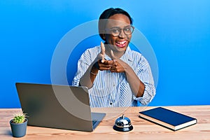 Young african woman working at hotel reception using laptop pointing fingers to camera with happy and funny face