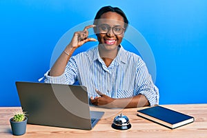 Young african woman working at hotel reception using laptop smiling and confident gesturing with hand doing small size sign with