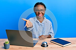 Young african woman working at hotel reception using laptop smiling friendly offering handshake as greeting and welcoming