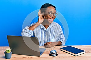 Young african woman working at hotel reception using laptop smiling with hand over ear listening an hearing to rumor or gossip