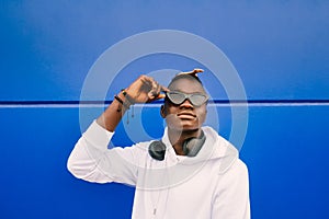 Young afro American black man wearing a white sweatshirt, sunglasses, comb and headphones on a blue wall with confidence