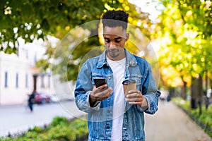 Young Afro American man with coffee cup standing smiling and use on cell phone in the city street