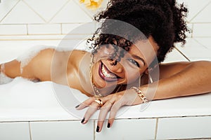 Young afro-american teen girl laying in bath with foam, wearing swag jewelry flawless, making selfie