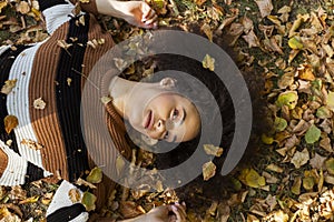 Young afro american woman lying on autumnal leaves in sunny park.