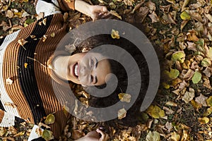 Young afro american woman lying on autumnal leaves in sunny park.