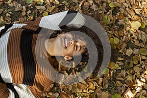 Young afro american woman lying on autumnal leaves in sunny park.
