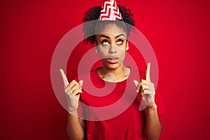 Young afro american woman wearing birthday hat over isolated red background amazed and surprised looking up and pointing with