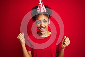 Young afro american woman wearing birthday hat over isolated red background celebrating surprised and amazed for success with arms