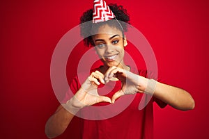 Young afro american woman wearing birthday hat over isolated red background smiling in love showing heart symbol and shape with