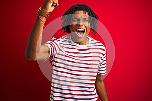 Young afro man with dreadlocks wearing striped t-shirt standing over isolated red background angry and mad raising fist frustrated