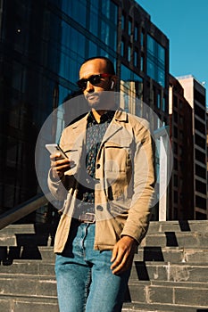 Young Afro man wearing sunglasses using smartphone to text message near modern business centre outdoor.