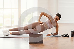 Young afro woman doing side plank on mat with pc