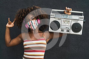Young Afro woman wearing face mask dancing outdoor while listening to music with wireless headphones and vintage boombox