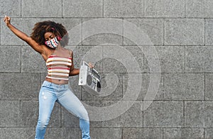 Young Afro woman wearing face mask dancing outdoor while listening to music with wireless headphones and vintage boombox