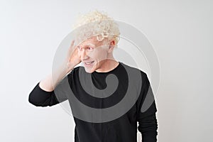 Young albino blond man wearing black t-shirt standing over isolated white background smiling with hand over ear listening an