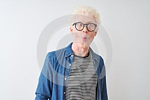 Young albino blond man wearing denim shirt and glasses over isolated white background making fish face with lips, crazy and
