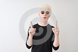 Young albino man wearing black t-shirt and sunglasess standing over isolated white background amazed and surprised looking up and