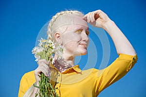 A young albino woman in a yellow blouse against a blue sky. holding a bouquet of daisies in her hands, in front of face