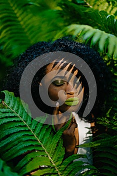 Young alluring african girl with green eyeshadows and lipstick is softly touching her face in the fern garden. Green