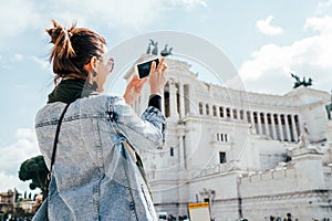 Young alone caucasian female tourist taking a photo of Victor Emmanuel II Monument using a modern smartphone on Piazza Venezia in