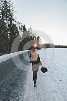 Young and alone girl dressed fur coat and dress standing around outdoor road. Sunset on the background. Sunlight and faded colors