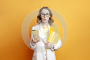 young amazed business girl in glasses and white suit holds documents and uses smartphone on colored background