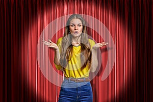 Young amazed, surprised girl wearing jeans and yellow shirt on red stage curtains background
