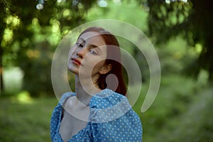 Young amazing woman in blue dress with red hair looking at the camera. The girl posing and enjoying walk in the woods.