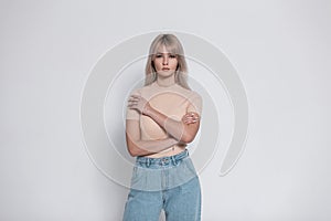 Young amazing woman in a fashionable shirt in vintage blue jeans stands near a modern white wall indoors. Pretty elegant girl