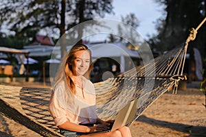 Young american girl using laptop and sitting on sand in wicker hammock.