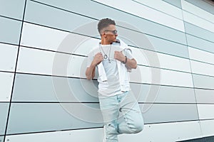 Young American hipster man in dark sunglasses in a fashionable white T-shirt in blue jeans relaxes near modern striped building in