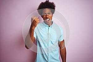 Young american man with afro hair wearing blue shirt standing over isolated pink background angry and mad raising fist frustrated
