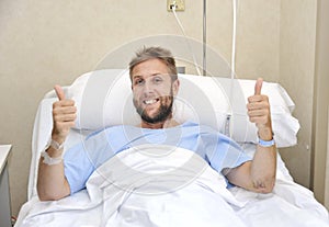 Young American man lying in bed at hospital room sick or ill but giving thumbs up smiling happy and positive